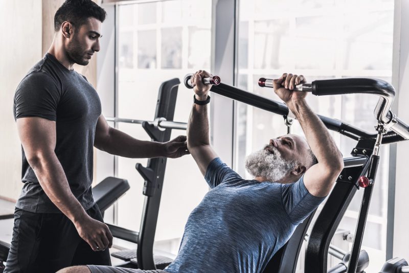 Senior bearded man is sitting at power training apparatus and hardly doing exercise in modern gym against big window. His instructor is standing nearby and watching him